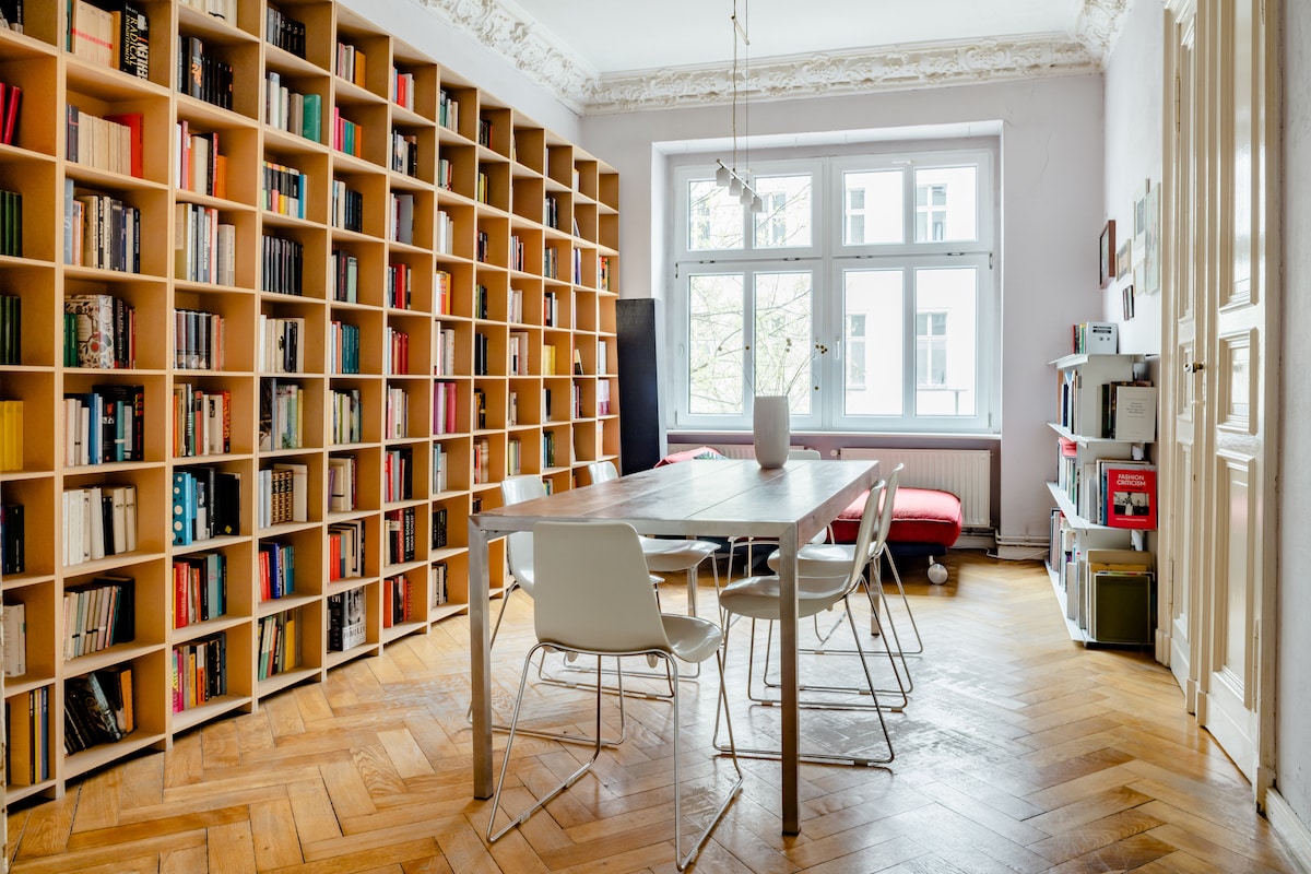 A bright room features a large bookshelf filled with colorful books along one wall. A table with white chairs occupies the center, while a cozy sleeping area with a red bed is visible in the background. Natural light flows through the large windows adorned with light curtains.