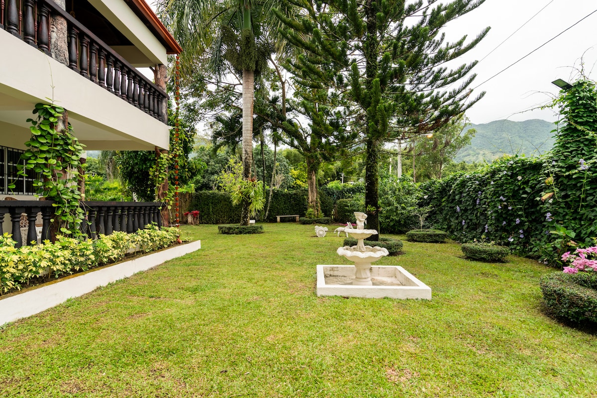 A spacious garden area is presented, framed by lush greenery and mountains in the background. A decorative fountain stands on a white base, surrounded by manicured grass and neatly arranged shrubbery. The villa's balconies are visible, enhancing the serene atmosphere.