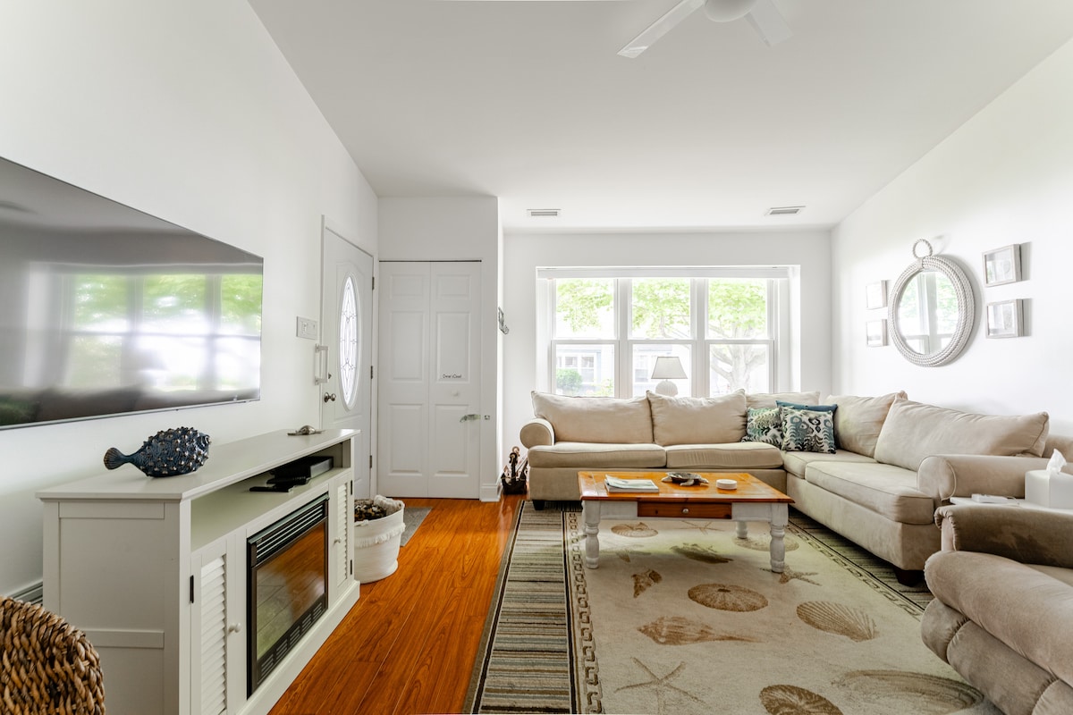 A bright living area is featured with ample natural light coming through large windows. A comfortable sectional sofa surrounds a low coffee table, while a decorative television stand adds visual interest. Light wooden flooring complements the relaxing ambiance of the space.