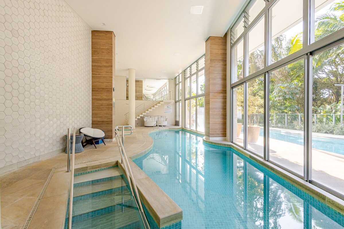 A serene indoor pool area features large windows that let in natural light, creating a bright atmosphere. The pool is outlined by pale blue tiles and has a gently sloping entry. Comfortable lounge chairs are placed nearby, offering relaxation options for guests.
