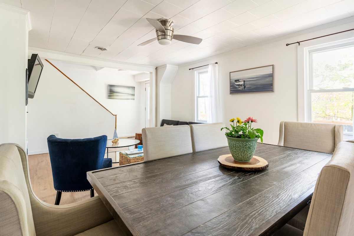 An open dining area features a large, rustic wooden table surrounded by soft, upholstered chairs. Natural light filters through the windows, highlighting a potted plant at the center. In the background, a cozy seating area includes a blue armchair and decorative artwork on the walls.
