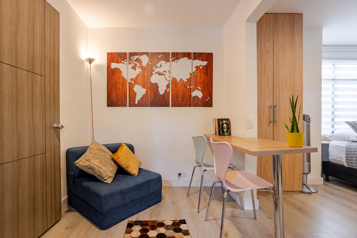 A compact sitting area features a blue upholstered chair topped with decorative cushions. A wooden table with colorful chairs is positioned nearby, while a wall art piece depicting a world map adds visual interest. Natural light fills the room, enhancing the neutral color scheme.