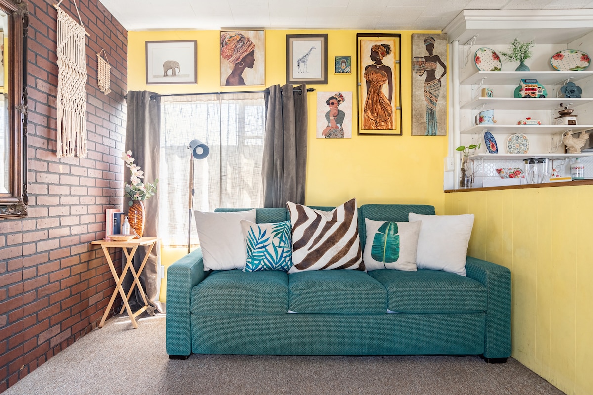 A bright living area features a teal sofa adorned with decorative pillows in various patterns. Colorful artwork is displayed on the yellow walls, while a wooden shelf showcases a collection of decorative items. Natural light filters through sheer curtains, enhancing the cozy ambiance.