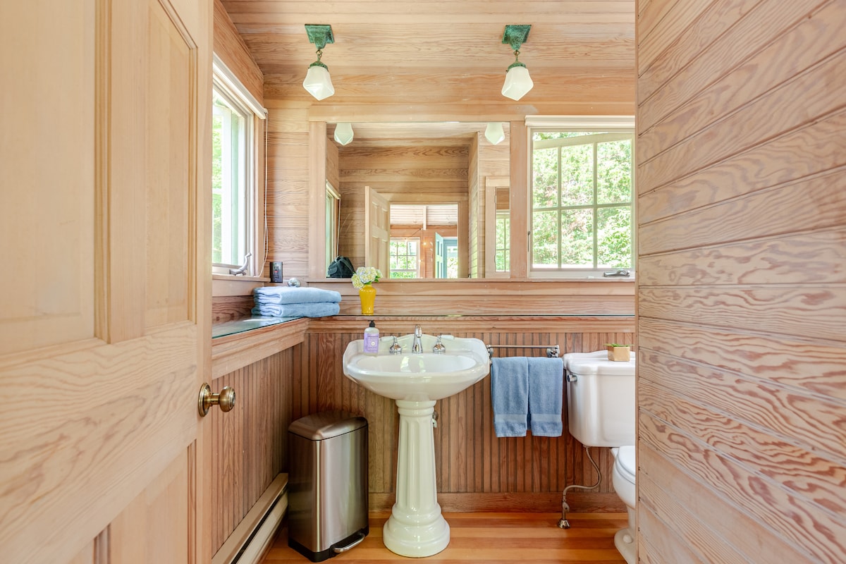 The spacious bathroom features wooden paneling and natural light streaming through multiple windows. A pedestal sink is complemented by a large mirror and modern light fixtures. Towel racks with fresh towels are visible, along with a waste bin and a toilet area, creating a functional layout.
