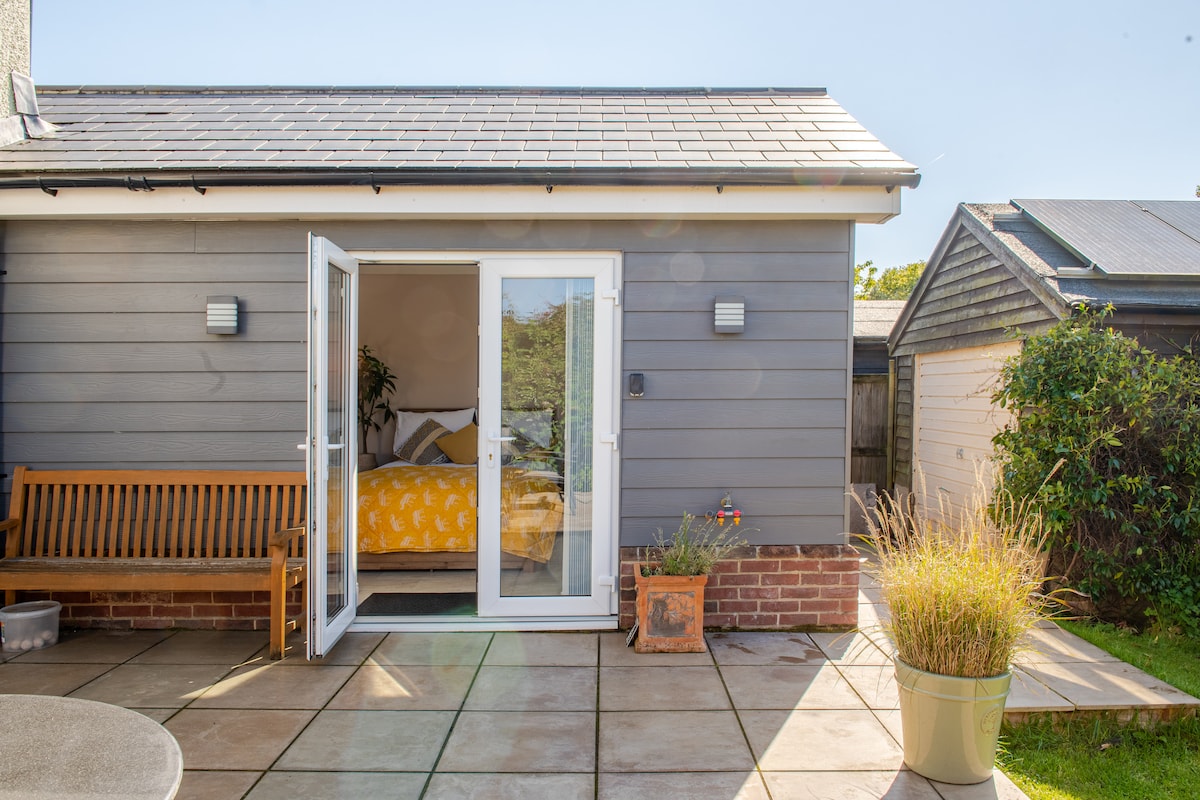 A private entrance is shown leading to a spacious room. Large glass doors provide direct access to a tiled patio area, featuring a wooden bench and potted plants. The muted color of the exterior complements the inviting surroundings.