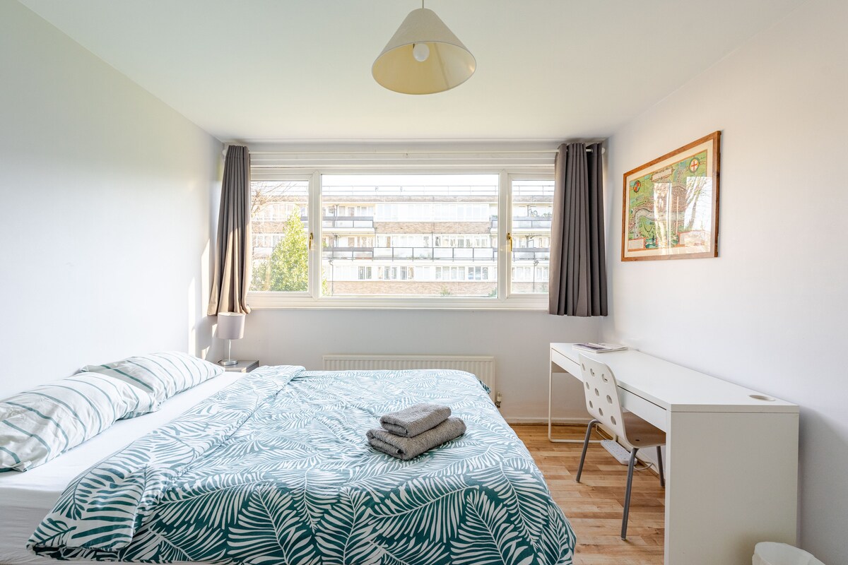 A bright and airy bedroom features a spacious bed adorned with a leafy patterned duvet. A large window allows natural light to fill the room, complemented by light-colored walls. A functional desk with a chair is positioned nearby, while neatly folded towels are placed on the bed.