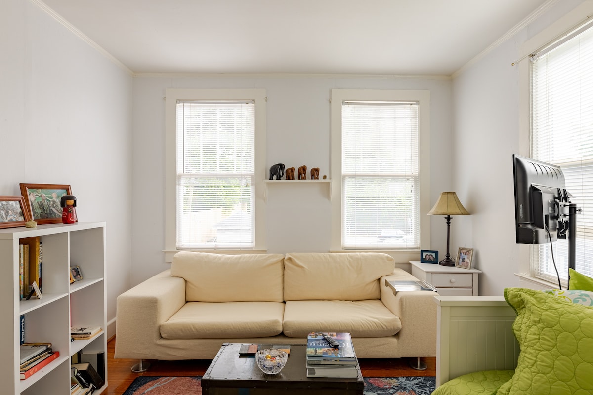 A bright living room is featured, showcasing a light-colored sofa positioned adjacent to two windows that allow ample natural light. A coffee table is centered in front of the sofa, and a small bookshelf is visible to one side, displaying various books and decorative items.