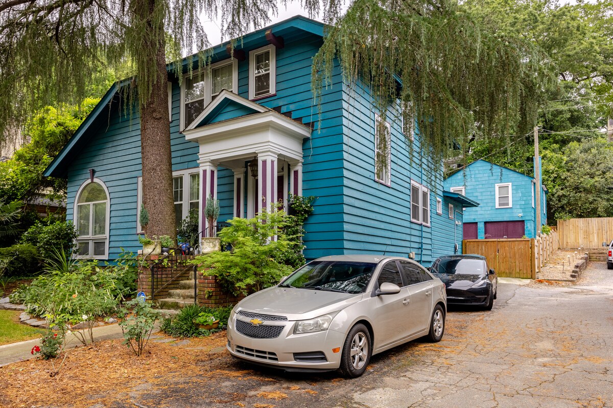 A charming blue house is set among greenery, showcasing a traditional architecture style. Two vehicles are parked in front, and the entrance is framed by steps leading to a porch adorned with potted plants. The surrounding area features a landscaped yard with foliage.