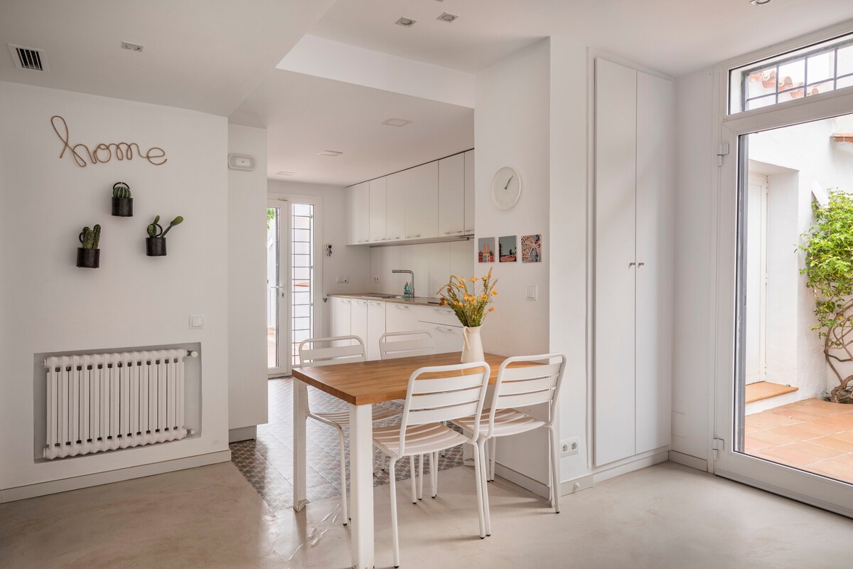 A modern kitchen and dining area feature a light wooden table surrounded by white chairs. The space is adorned with minimalistic decor, including potted plants on the wall. Natural light flows through large windows, highlighting the clean lines and open layout.