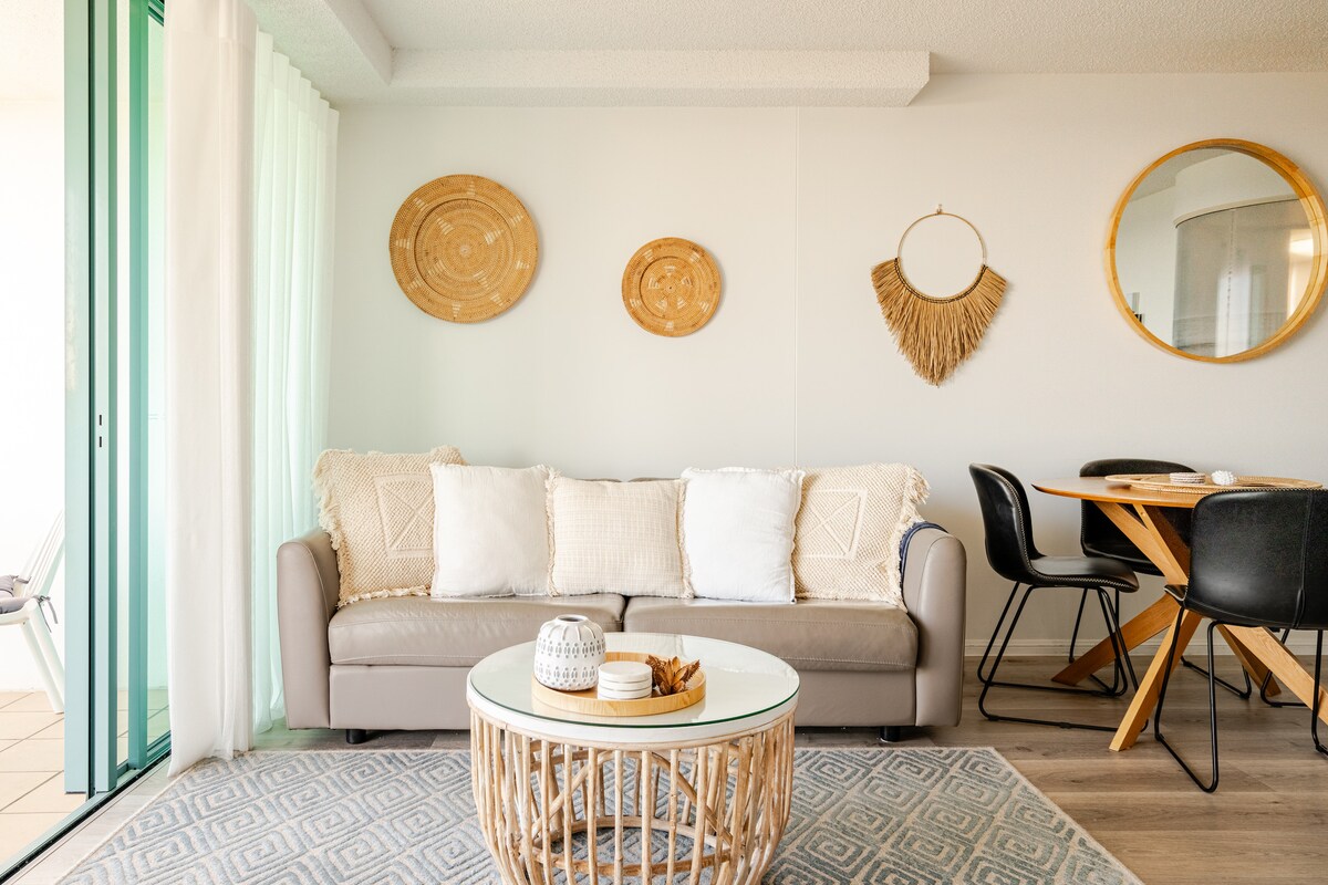 A cozy living area features a tan sofa adorned with light cushions, complemented by a round coffee table. Decorative wall plates and a woven wall hanging add texture, while a large mirror enhances the space. Nearby, a dining table with black chairs is visible.