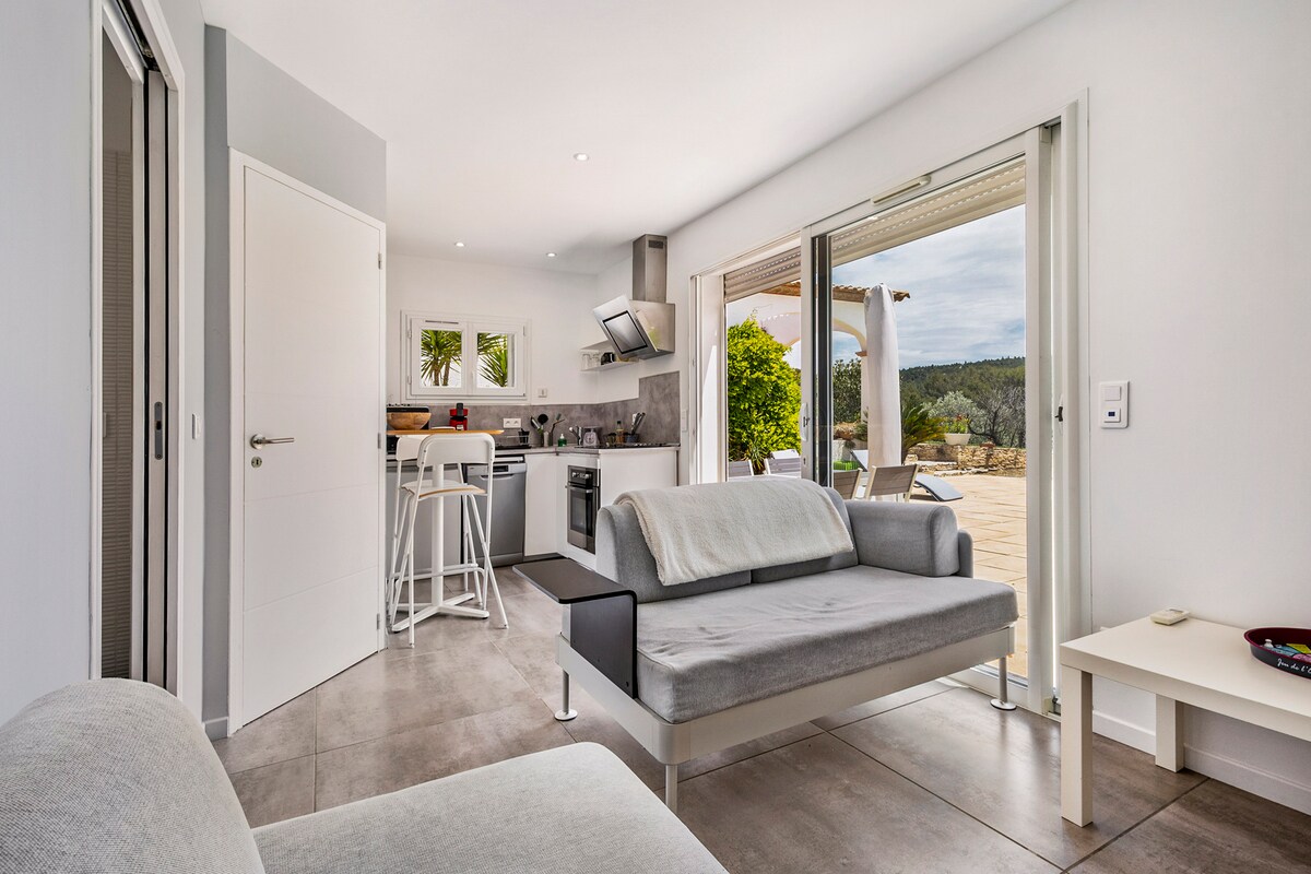 An open-plan living area features a modern kitchen equipped with essential appliances, including a dishwasher and a microwave. A gray sofa is positioned nearby, with glass doors that open to a patio overlooking the scenic outdoors.
