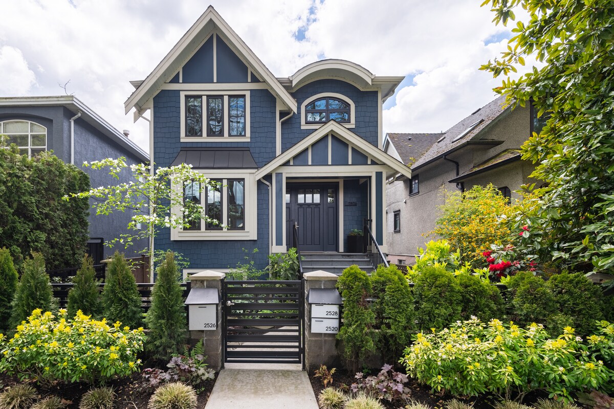 A charming blue house is framed by lush greenery and colorful flower beds. The entrance features a modern black metal gate leading to a pathway, with large windows allowing natural light to enhance the exterior design.