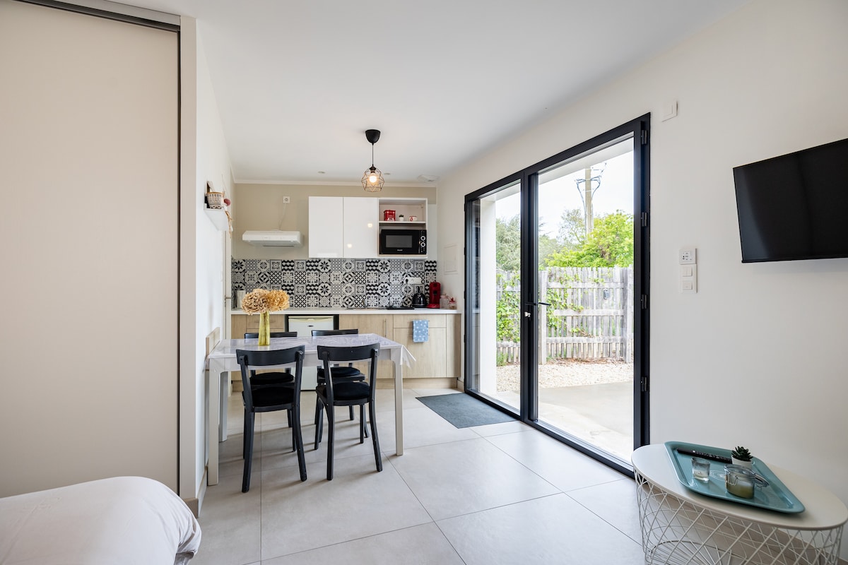 An open-plan living area is captured, featuring a modern kitchenette with patterned tiles. A dining table with four chairs is positioned nearby. Large glass doors provide access to the outdoor area, allowing natural light to brighten the space. A wall-mounted TV is visible.