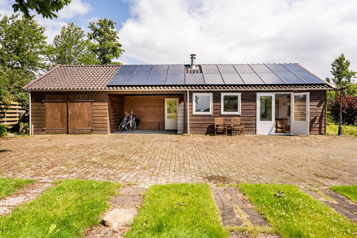 A rural vacation home is shown, featuring a sloped roof equipped with solar panels. A well-maintained stone patio provides access to the entrance, where wooden chairs and a table are placed. Two bicycles are visible nearby, surrounded by greenery and trees.