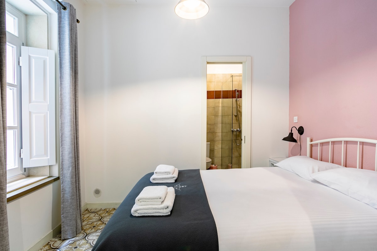 A well-lit sleeping area features a spacious bed, neatly arranged with towels at the foot. Soft grey curtains frame a window, while a light fixture hangs above. An en-suite bathroom is partially visible through an open door, showcasing tasteful tilework.