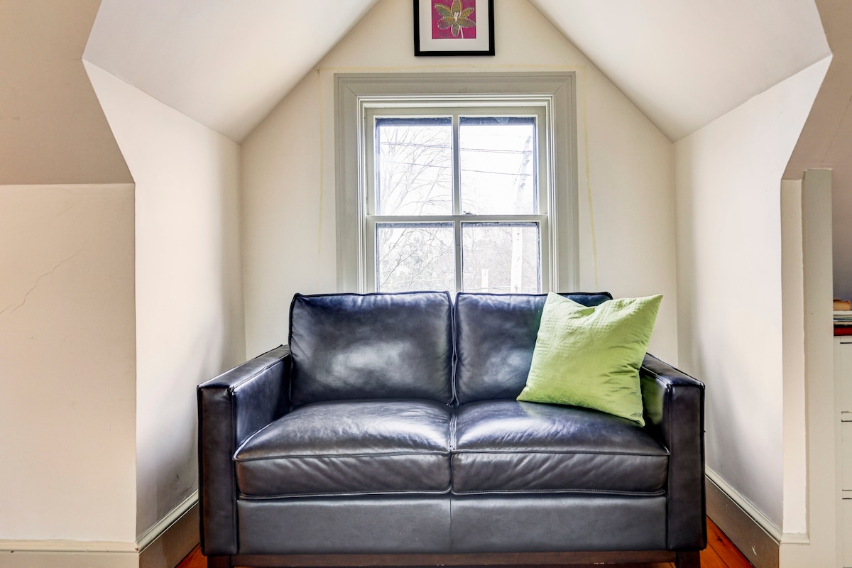 A cozy futon couch is positioned beneath a sloped ceiling, featuring a green accent pillow. Natural light enters through a window, enhancing the room's spacious feel. A framed floral artwork adorns the wall above, contributing to the inviting ambiance.