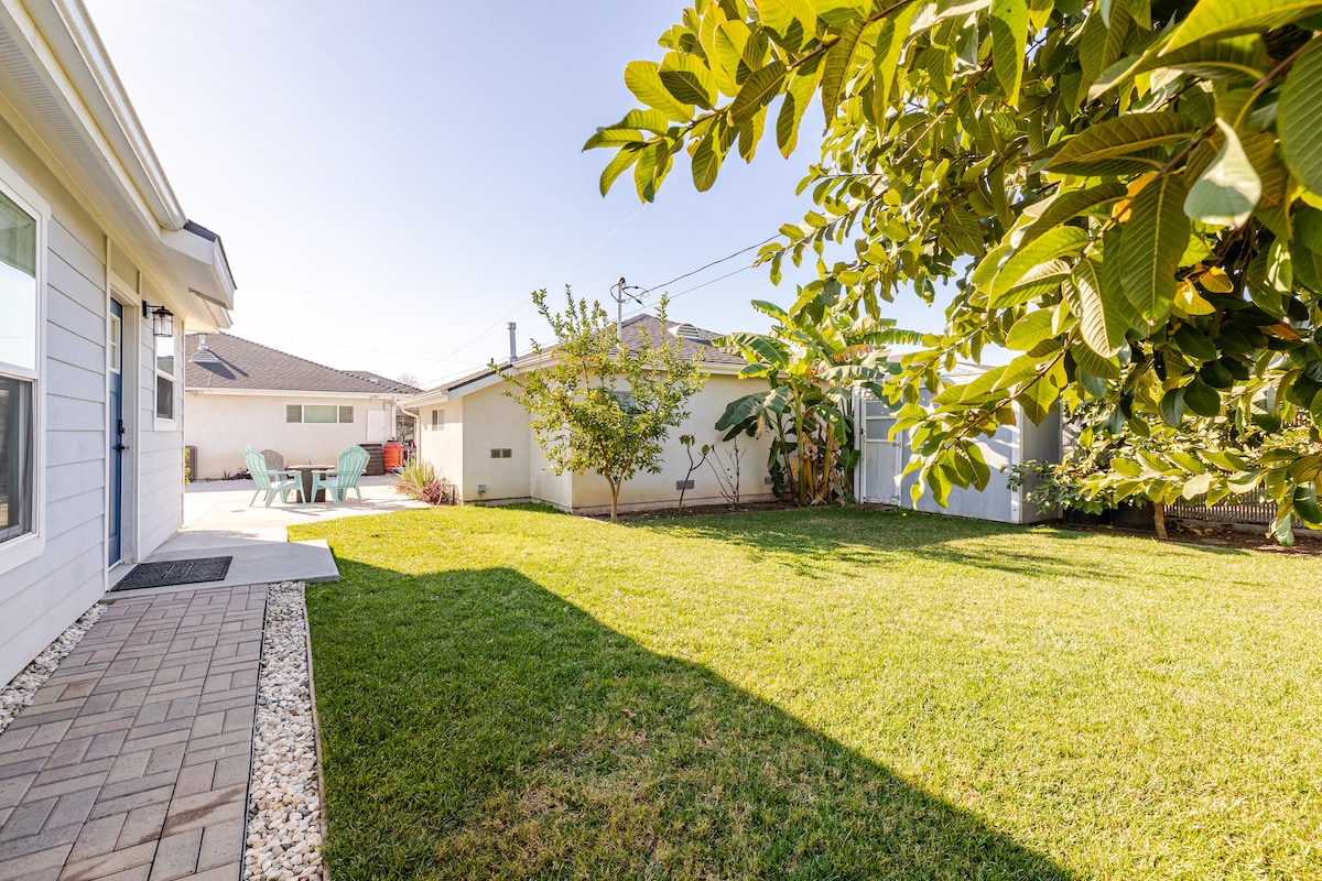 A spacious backyard is featured, showcasing well-maintained green grass under a bright sky. Small seating areas with chairs are visible, surrounded by various plants and trees, adding to the inviting outdoor space. The patio area offers an opportunity for relaxation or outdoor activities.