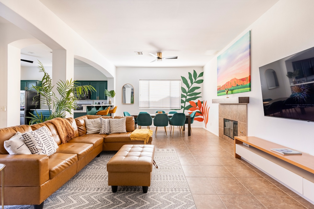 A spacious living room features a brown leather sectional sofa and a decorative ottoman. The area is complemented by a large wall-mounted flat-screen TV and a fireplace. Colorful art adorns the walls, and a dining area is visible in the background, showcasing a large table surrounded by chairs.
