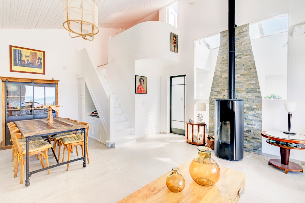 A bright living area is presented with large windows allowing natural light. A wooden dining table with rattan chairs is centrally placed, while a sleek black fireplace stands against a stone accent wall. Art pieces adorn the walls, and a staircase leads to the upper level.