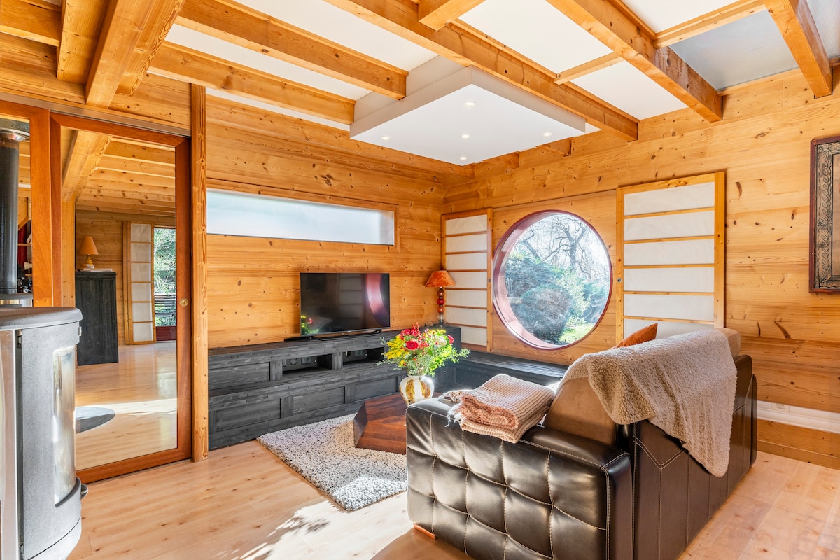 A warm and inviting living area features wood paneling and natural light from a circular window. A comfortable sofa is positioned beside a dark media unit beneath a wall-mounted television. A coffee table with a decorative arrangement is placed in the center, enhancing the cozy ambiance.