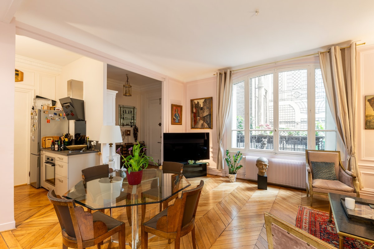 A well-lit living area showcases a round dining table surrounded by elegant chairs. A small kitchen area is visible to the left, with modern appliances. A large window features natural light, and potted plants add a touch of greenery, enhancing the apartment's inviting ambiance.