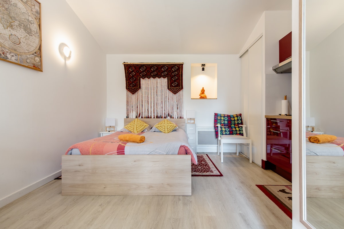 A welcoming studio space features a spacious bed adorned with decorative pillows and a colorful blanket. A small kitchenette area is visible, along with a cozy chair positioned beside an accent wall. Light is softly illuminated across the room, enhancing its calm ambiance.