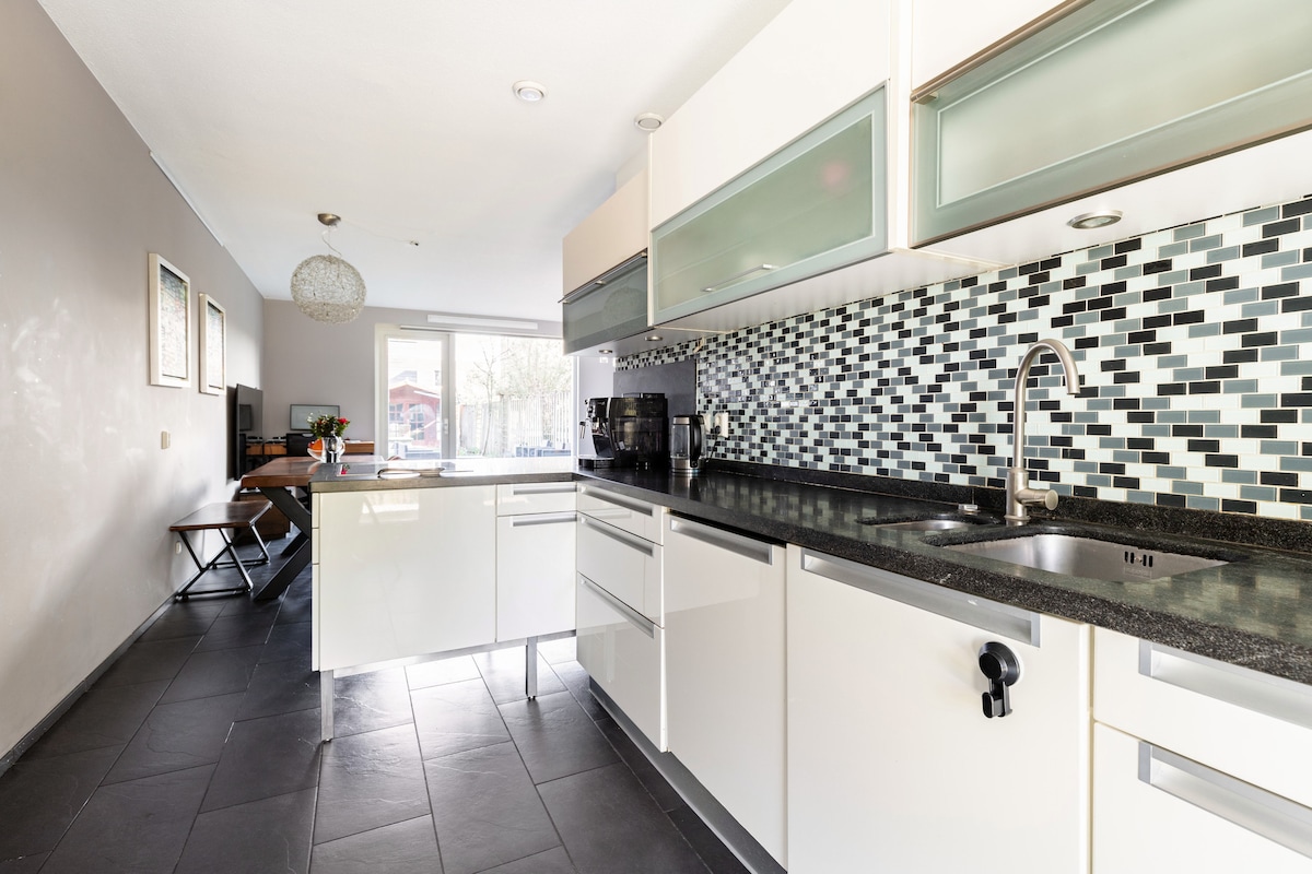 A modern kitchen is shown, featuring sleek white cabinetry and a striking mosaic backsplash. A blend of natural light and overhead fixtures illuminates the space. A dining area is visible with a minimalist table and chair setup, enhancing a functional layout.