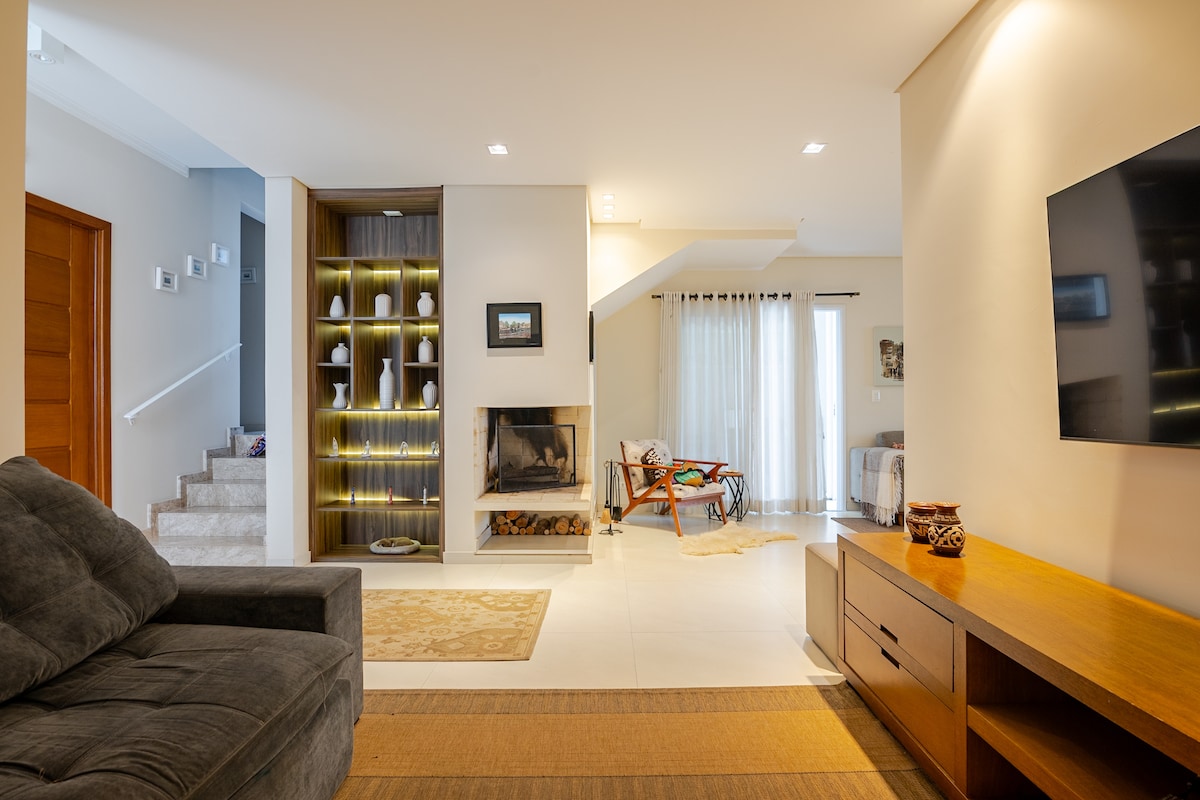 The spacious living area features a comfortable gray sofa and a wooden media console with a television. Soft lighting highlights decorative items in built-in shelving. A staircase is visible in the background, leading to the upper level, while large windows allow natural light to fill the room.