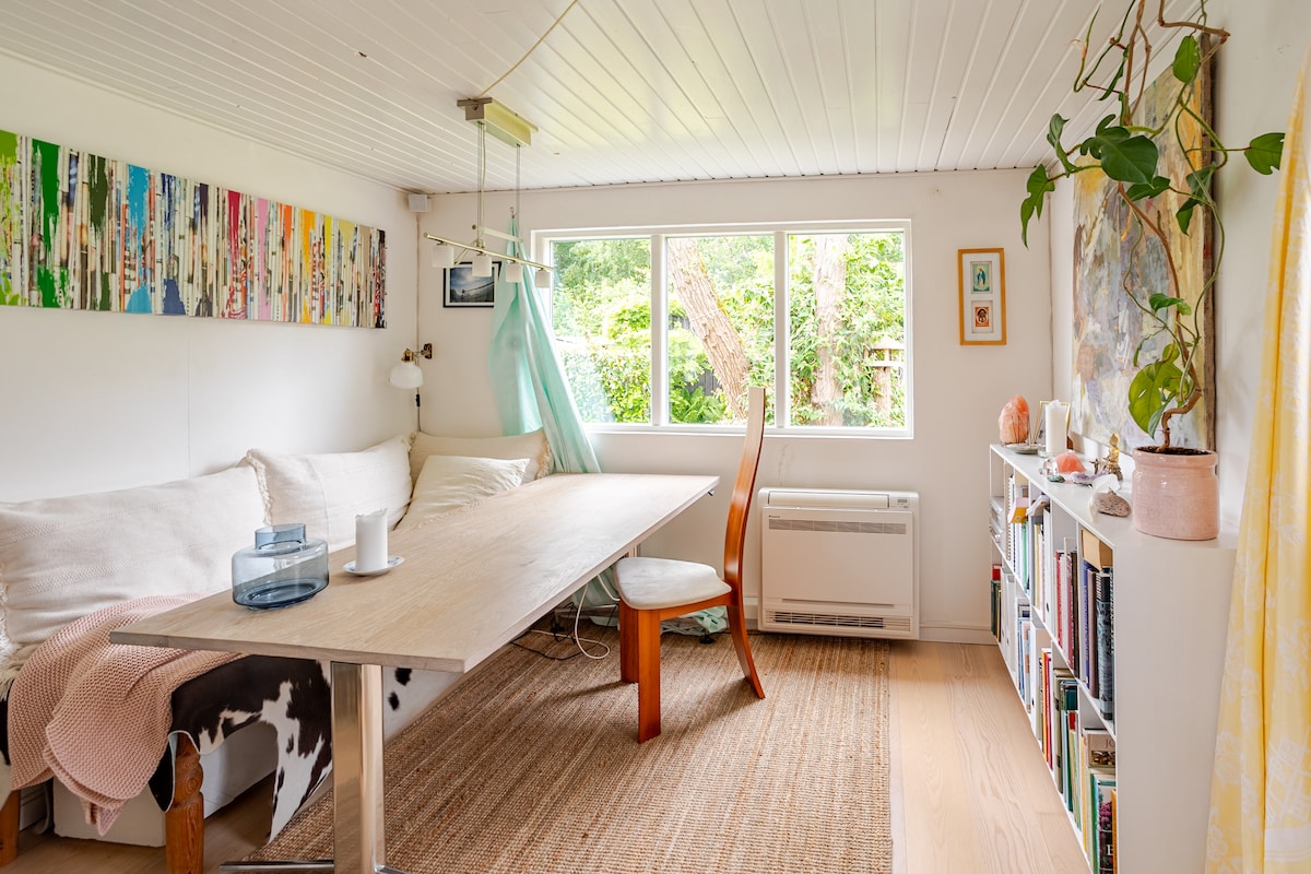 A light-filled interior features a large wooden table surrounded by an orange chair and white accents. A comfortable sofa is positioned nearby, with colorful artwork displayed on the walls. Natural greenery is visible through the windows, enhancing the serene environment.