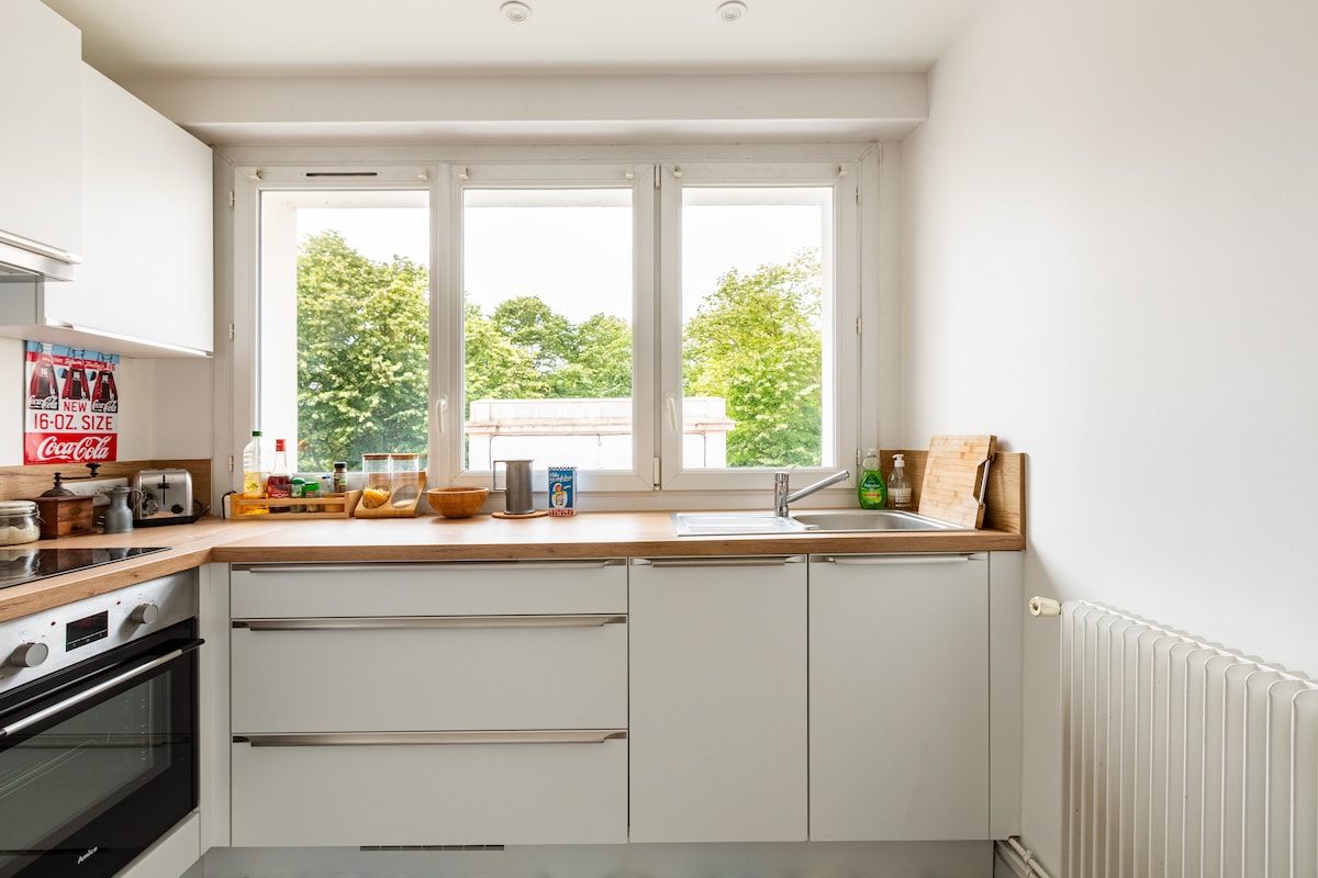 A functional kitchen features light-colored cabinetry with a wooden countertop. Natural light filters through three windows, showcasing a view of greenery outside. Appliances include an oven and stovetop, with kitchen essentials neatly arranged, creating an organized and inviting space.