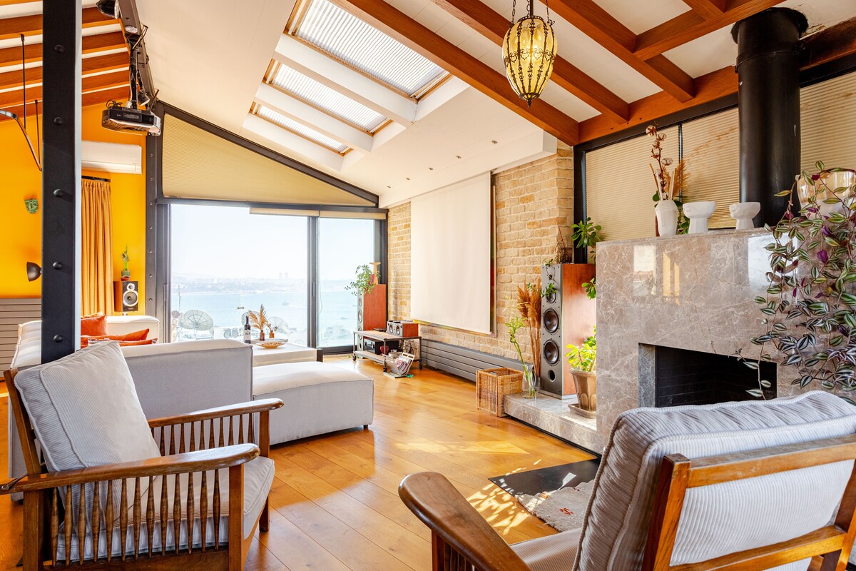 A spacious living area features large windows that provide views of the bay. Natural light fills the room, highlighting the wooden ceiling beams. Comfortable seating includes two armchairs and a sofa, accompanied by a modern fireplace and decorative elements on the mantle.
