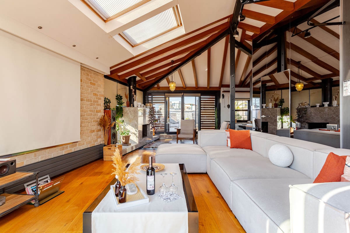 An open living area features a spacious sectional sofa with decorative cushions, complemented by a coffee table that holds a bottle of wine and glasses. Tall windows allow natural light to fill the space, accentuating the wooden beams and warm tones of the floor.