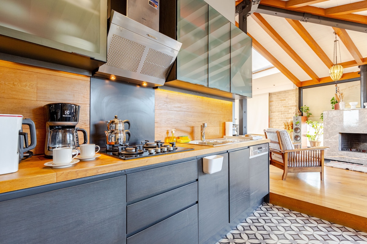 The kitchen area features sleek cabinetry and a polished wooden countertop. A gas stove is positioned alongside a coffee maker and kettle. Under-cabinet lighting highlights the workspace, while decorative tiles provide texture to the flooring.