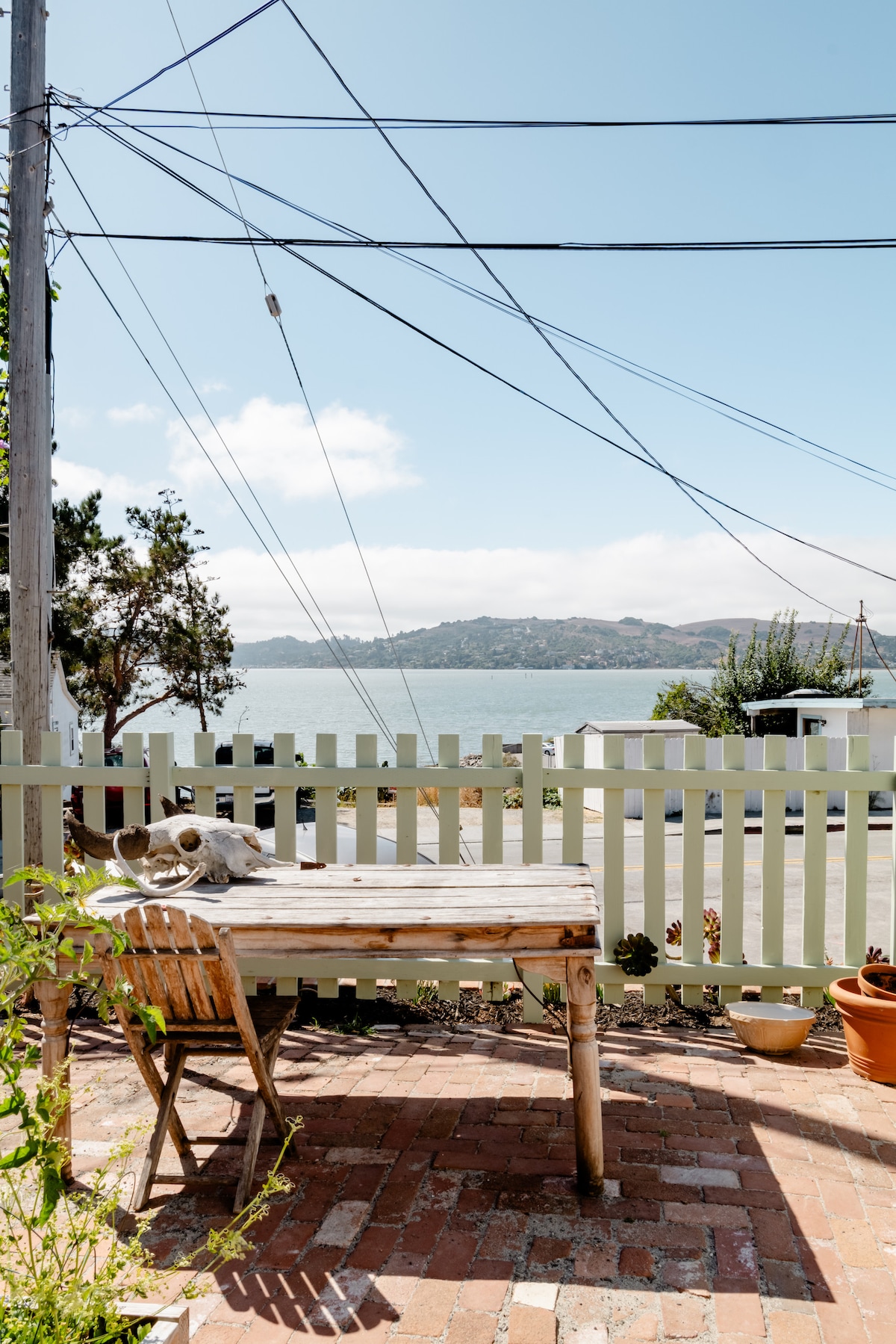 A serene patio area features a wooden table and chair set against a fence, with views of the bay and distant hills. Natural light illuminates the space, enhancing the quiet retreat setting. Potted plants add a subtle touch of greenery.