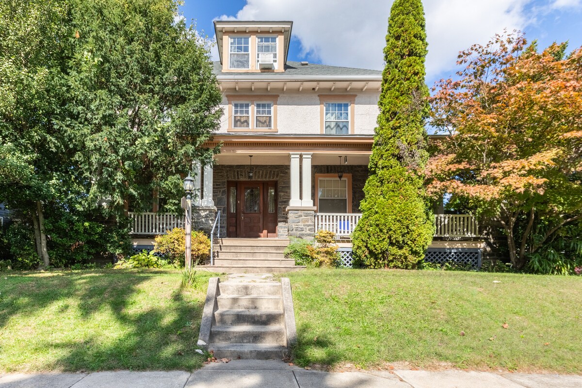 The exterior of the home features a well-maintained façade with a blend of stone and siding, complemented by a welcoming porch and double front doors. Lush greenery, including a tall evergreen and colorful foliage, accents the inviting entrance and well-kept front yard.