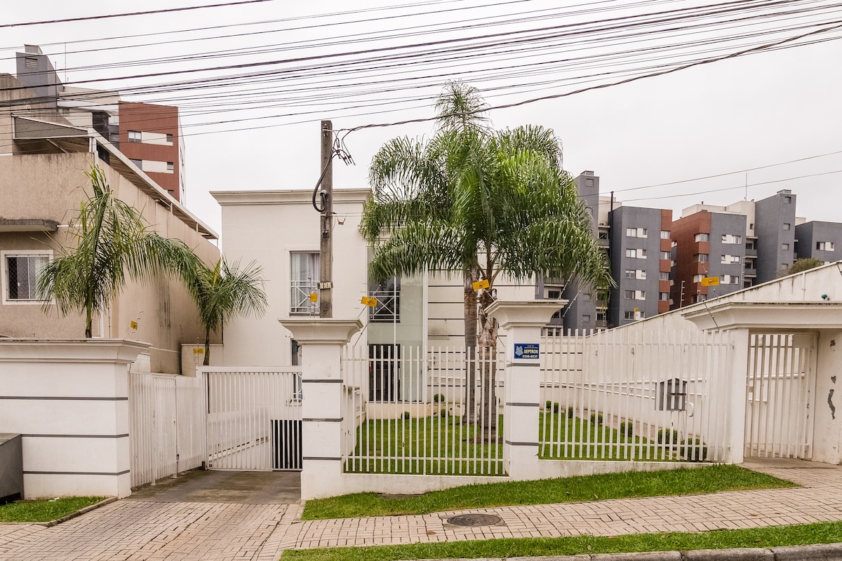 A modern residential building is set behind a white picket fence, featuring manicured green lawns and palm trees. The structure is flanked by nearby urban buildings, offering a blend of greenery and cityscape. The entrance is marked by a gate with surrounding concrete pathways.