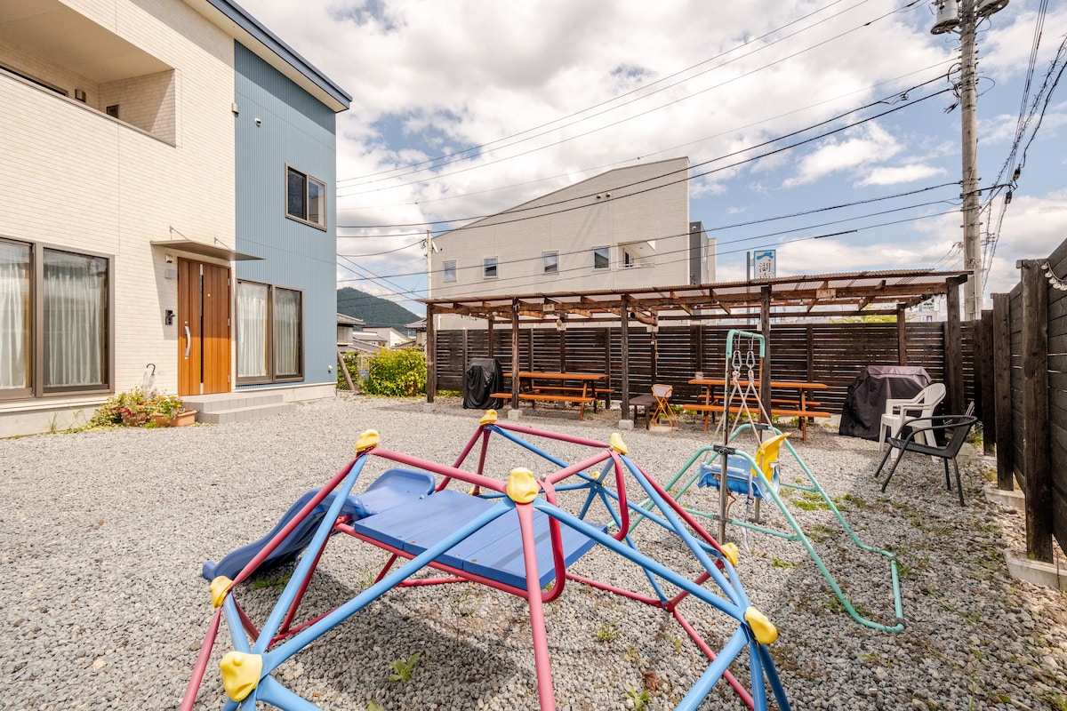 An outdoor space featuring a gravel ground with a colorful play structure and seating area. Several picnic tables are sheltered under a wooden canopy, providing a gathering space. Nearby, cooking facilities are visible, enhancing the area's functionality for outdoor activities.