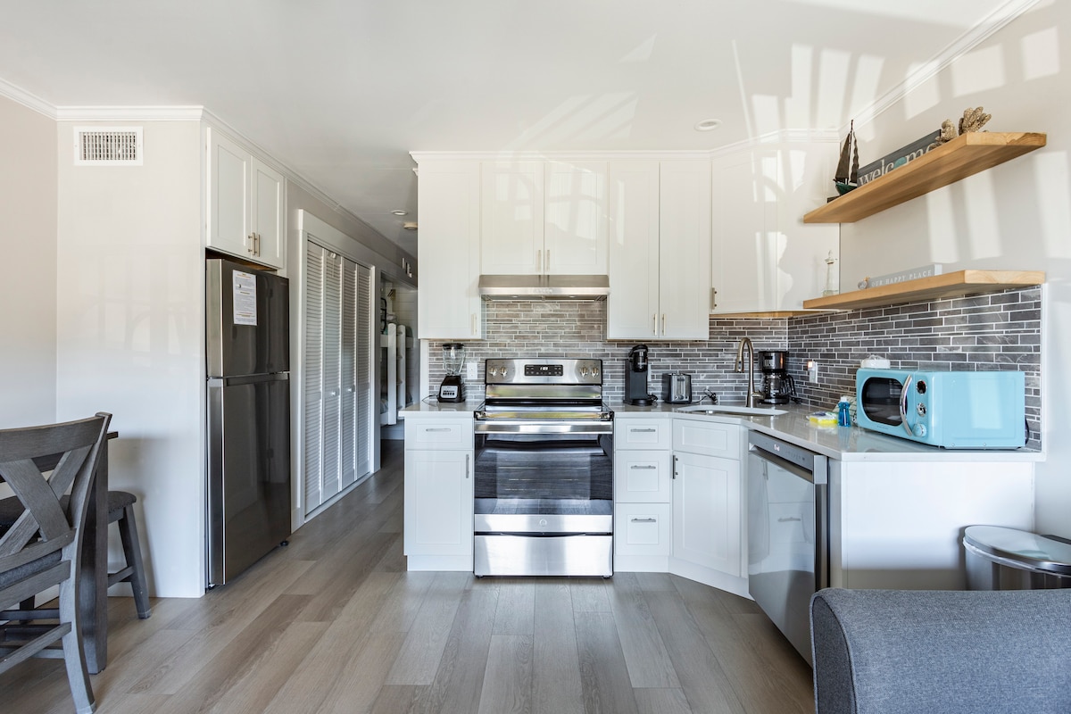 A modern kitchen is equipped with stainless steel appliances, including a refrigerator, stove, and microwave. Light-colored cabinetry and a tiled backsplash complement the design, while wooden shelves hold decorative items. A small dining area with chairs is visible, enhancing the kitchen's functionality.