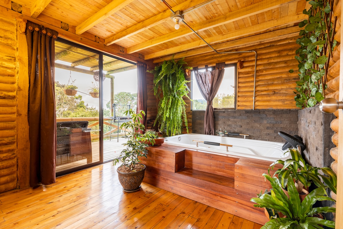A wooden interior bathroom features a large, inviting hot tub surrounded by natural elements. Plants are positioned nearby, adding greenery to the space. Large glass doors offer views of the outdoor area, while warm wooden beams and flooring create a cozy atmosphere.