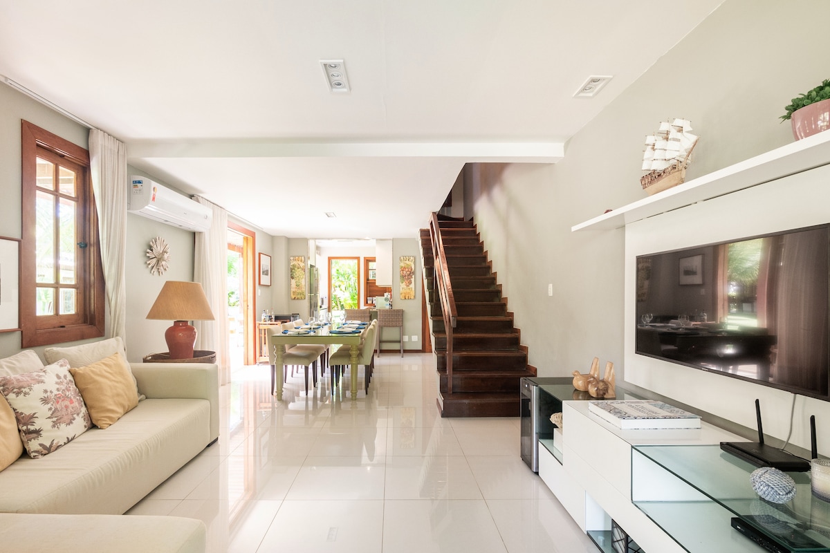 A well-lit living area features a comfortable seating arrangement with soft cushions. A staircase leads to the upper level while a dining space is visible in the background. Large windows provide natural light and views of the outdoor greenery.