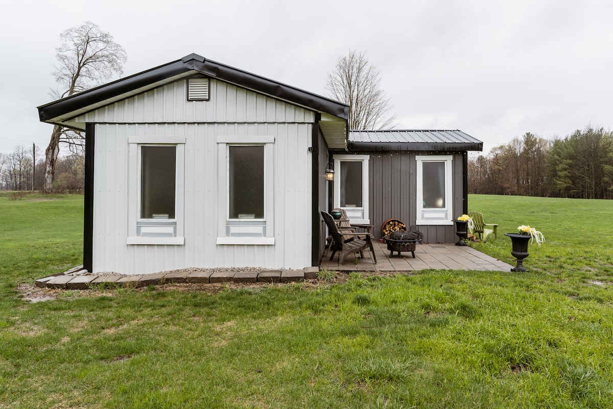 The exterior of the Bunky is shown, featuring a combination of white and dark grey siding. A porch area with wooden planks provides seating options. Green grass surrounds the building, set against a backdrop of trees in the distance under a cloudy sky.