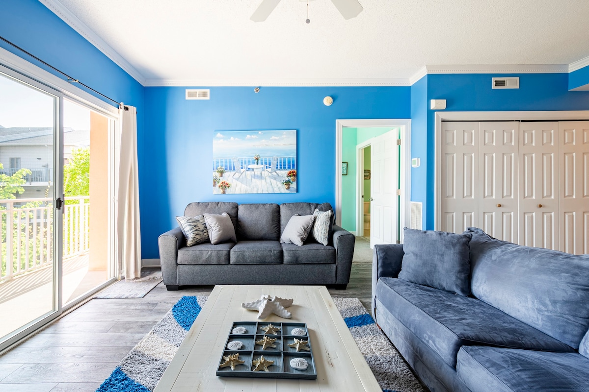 A bright living area features blue walls complemented by light-colored furniture. Two sofas are positioned around a centrally placed coffee table, which holds decorative items. Large glass sliding doors open to a balcony, allowing natural light to fill the space.