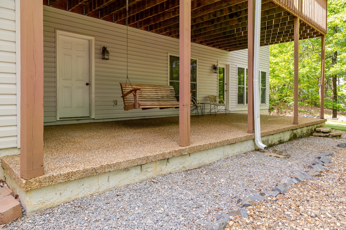 A covered patio area with a swing and a small table is seen under a wooden deck. The surface is made of a textured finish, surrounded by gravel and a landscaped area. Light filters through the trees in the background, providing a natural setting.