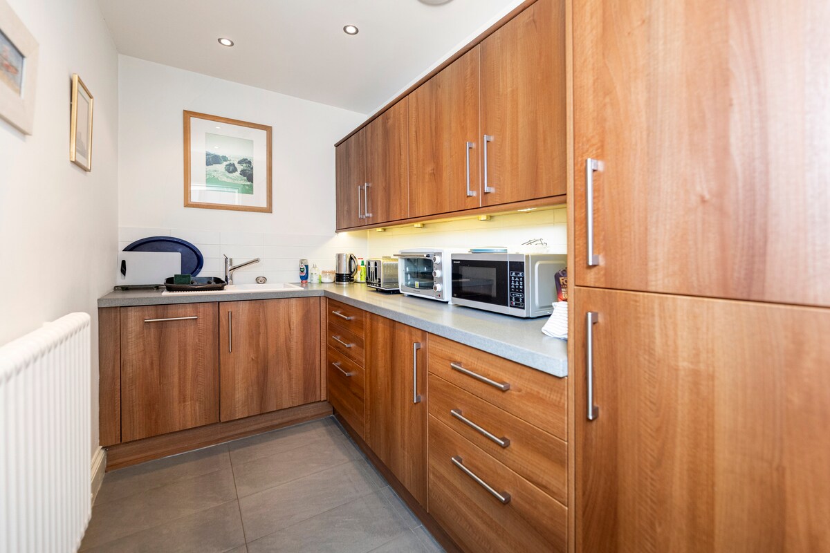A functional kitchen area features warm wooden cabinetry complemented by a gray countertop. Various modern appliances, including a microwave, toaster, and kettle, are neatly arranged, providing convenience and utility. Soft lighting enhances the space while a framed picture adds a touch of decor.