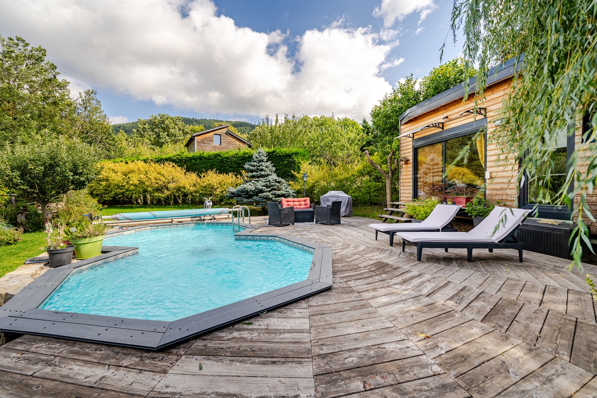 An inviting outdoor area features a private pool surrounded by a wooden deck. Two lounge chairs are positioned nearby, and lush greenery creates a natural backdrop. A seating arrangement is visible, along with a view of the pool house through large glass doors.