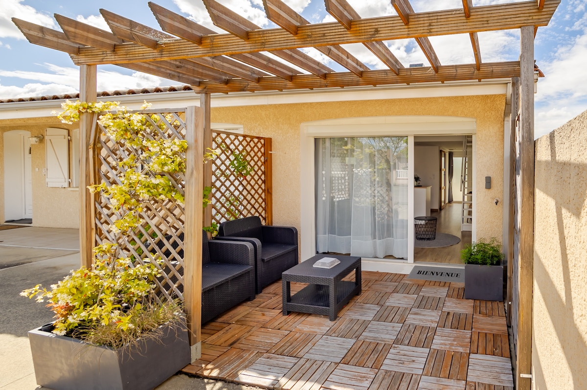 An outdoor seating area is featured beneath a wooden pergola, showcasing a dark seating set and a low table. The textured floor is made of wooden tiles and is surrounded by green plants, creating a serene entrance that leads to the interior.