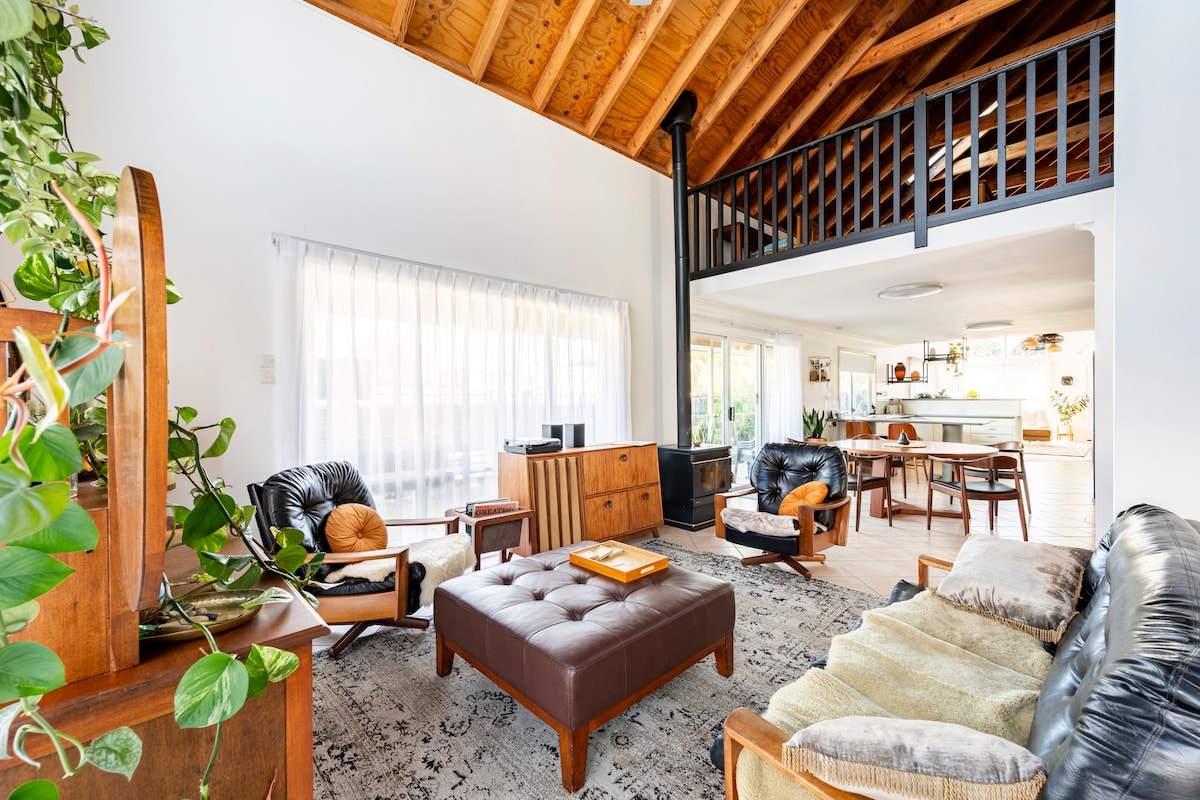 A spacious living area features high wooden ceilings and an open layout. Comfortable seating arrangements include stylish leather chairs and a large ottoman, complemented by greenery and natural light from large windows. A cozy fireplace is situated in the corner, enhancing the inviting space.