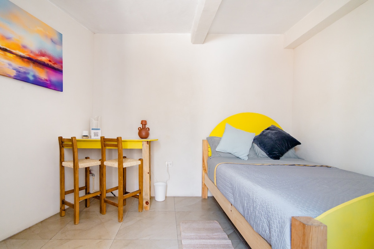 A bright room features a spacious bed with light grey bedding and colorful accent pillows. A small wooden table with two chairs is set in the corner, alongside a vibrant artwork that adds a splash of color to the white walls.