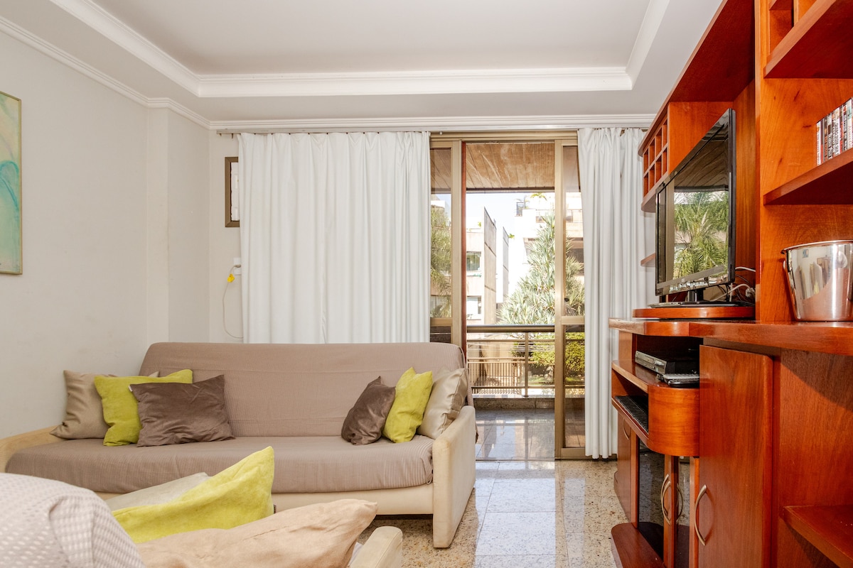 The living area features a spacious sofa with decorative cushions, complemented by a wooden media console. Floor-to-ceiling windows allow natural light to enter, offering views of the outdoors. Light-colored drapes frame the windows, enhancing the openness of the room.