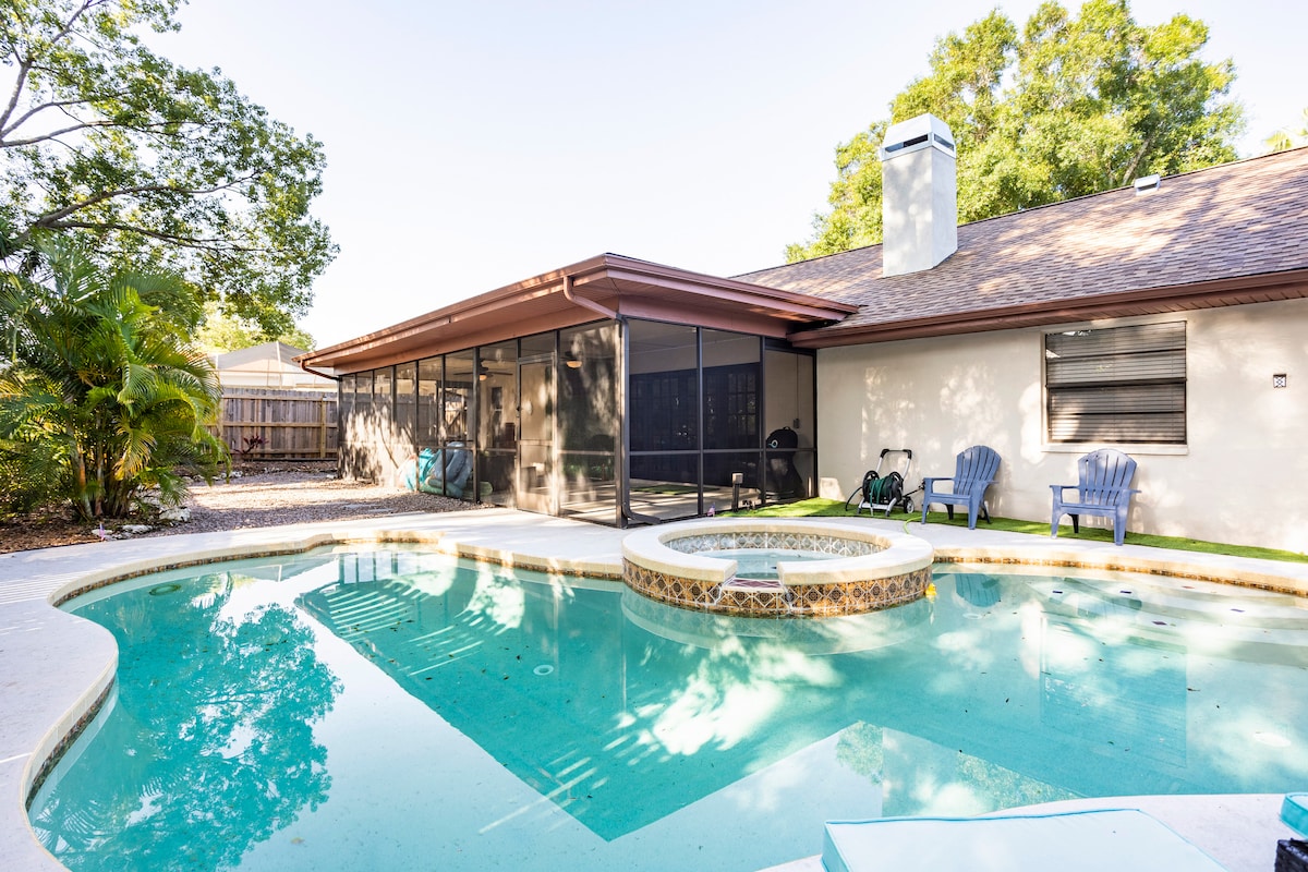 An inviting outdoor area features a screened-in patio adjacent to a swimming pool. The pool, with varying depths, includes an integrated jetted spa that overflows into it. Two lounge chairs are positioned nearby, surrounded by green foliage, contributing to a serene setting.